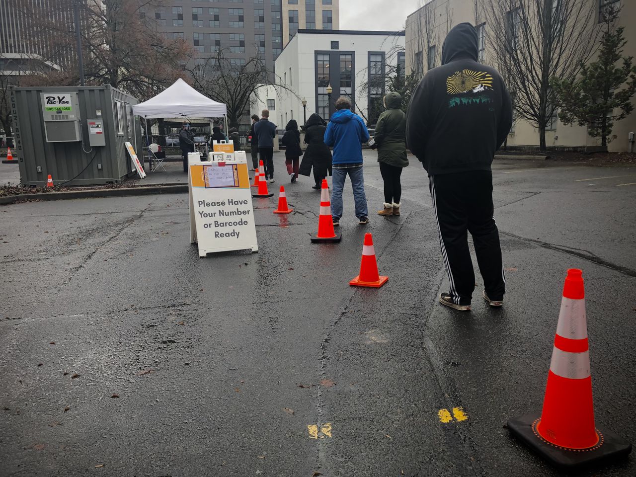 A line of people stand in a parking lot waiting to get tested for COVID-19. There are orange traffic cones lining up next to them and a white tent under which people are getting tested.