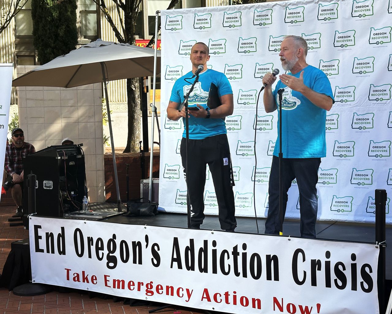 Two men stand on a temporary stage at Pioneer Square with a banner that says: "End Oregon's Addiction Crisis" and "Take Emergency Action Now!"