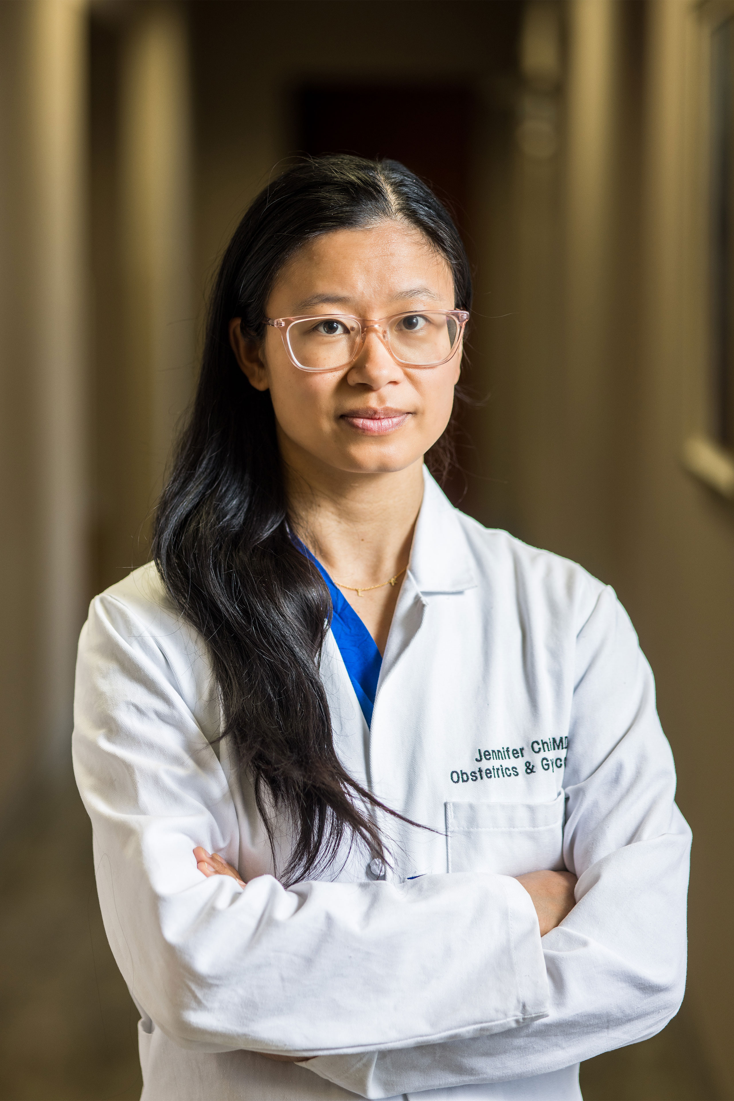 A woman with long dark hair and wearing glasses, dark blue scrubs, and a white doctor's coat stands with her arms crossed and looks at the camera.