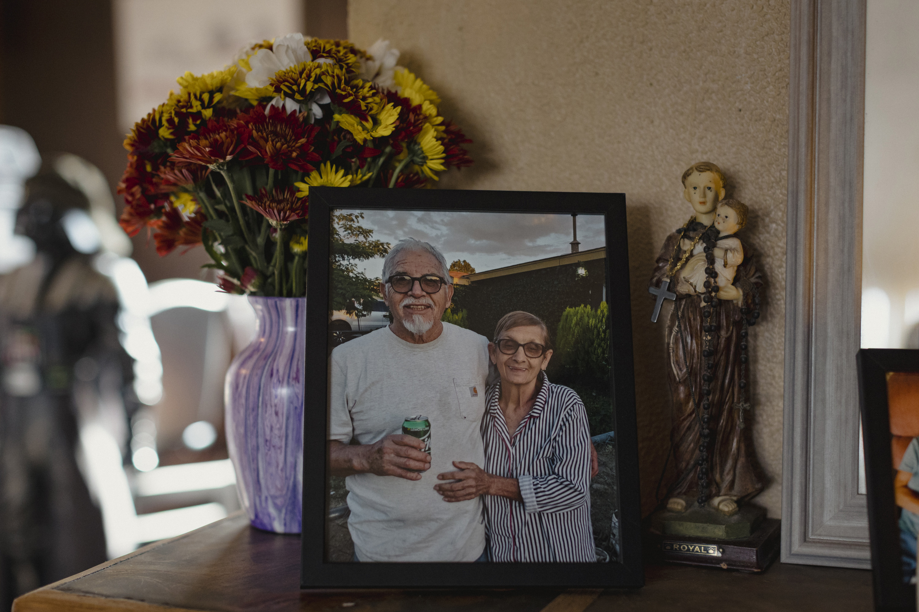 A framed photo of a man and woman sits on a tabletop between flowers and a religious figurine sculpture