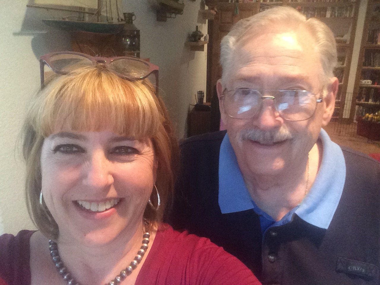 A woman wearing a red shirt takes a selfie of herself with a man wearing a blue shirt and glasses