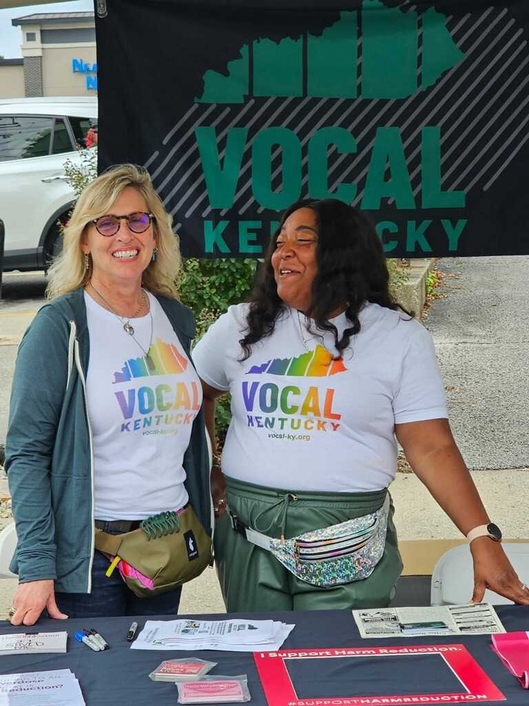 Two women wearing t-shirts reading Vocal Kentucky stand behind a table with with various papers and pens on it.